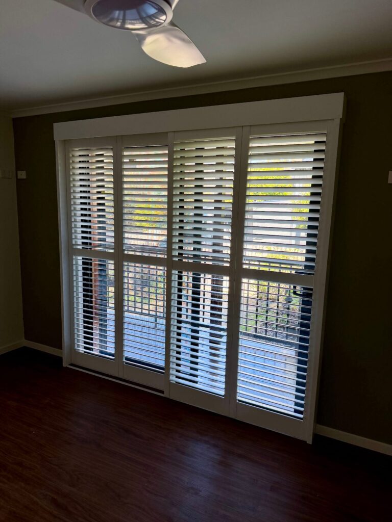 Four white PVC plantation shutter panels fitted across a full-height door opening in a Caboolture home, louvres tilted open above the mid rail